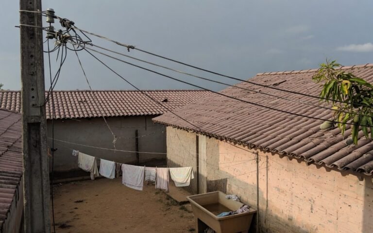 Cabos de energia passando na altura do telhado de uma casa simples, com poste de concreto envelhecido e fiacao irregular